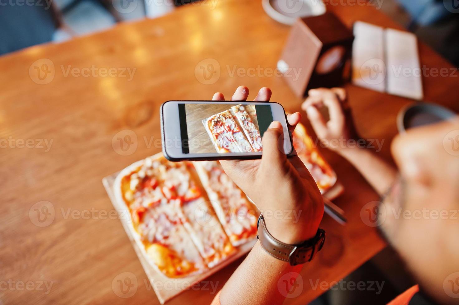 zelfverzekerde jonge indiase man in oranje trui zit bij pizzeria, eet pizza en maakt foto op zijn telefoon.