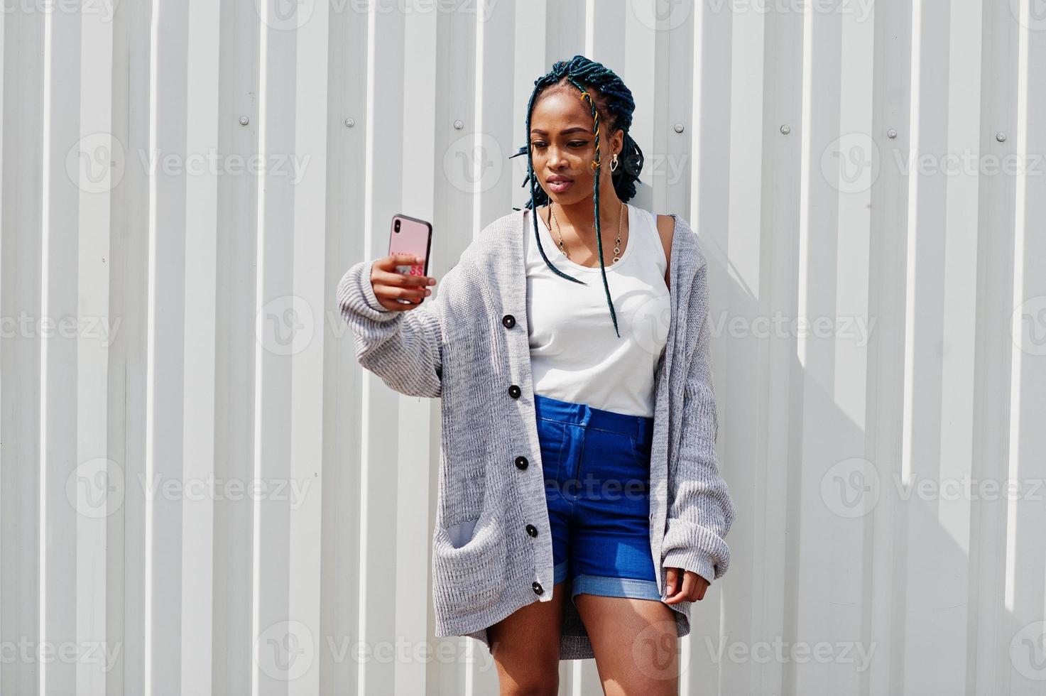 afrikaanse vrouw met dreadlocks haar, in spijkerbroek gesteld tegen witte stalen muur en selfie maken via de telefoon. foto