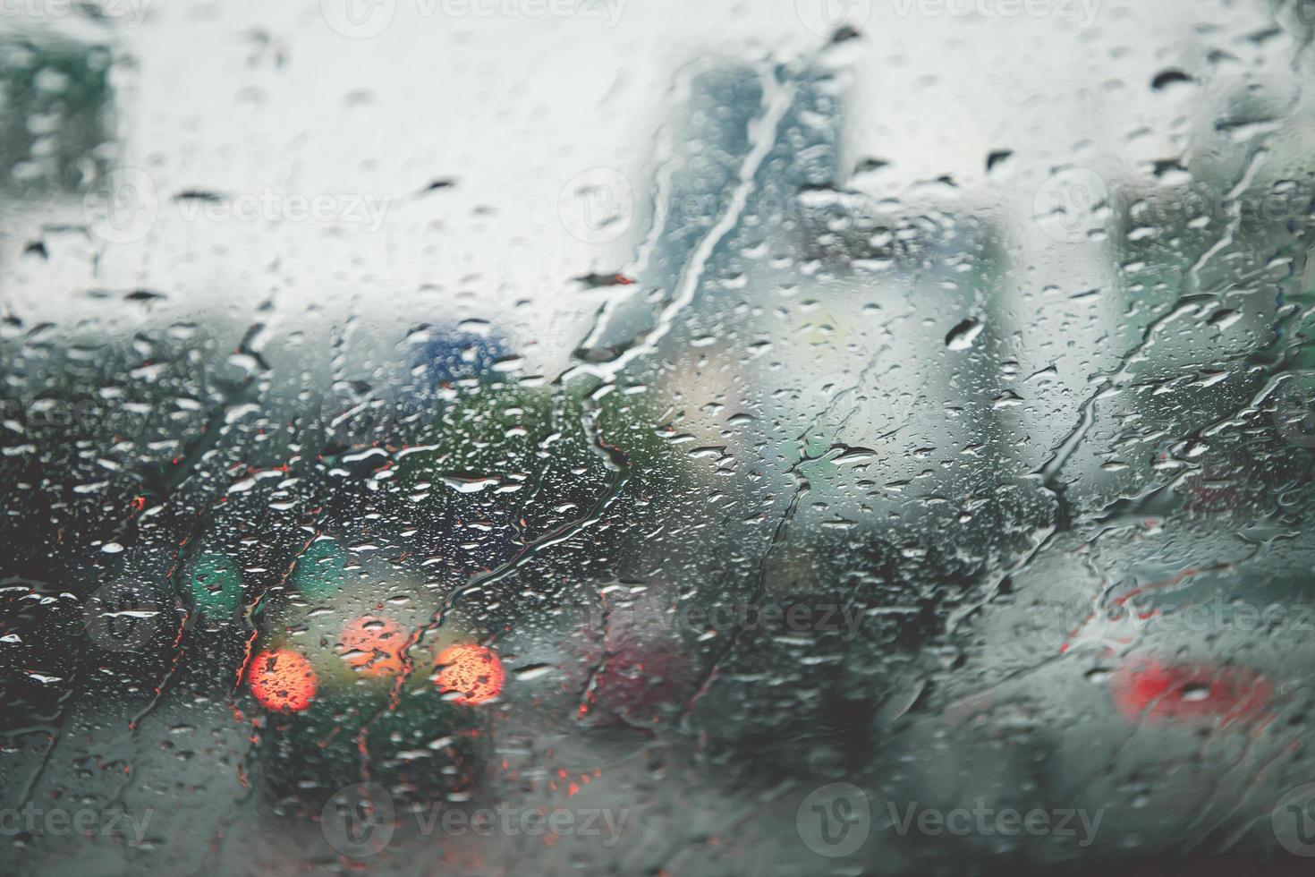 druppels regen motregen op de glazen voorruit in de avond. straat in de zware regen. bokeh achterlicht en verkeerslichten in de stad. rijd alstublieft voorzichtig met de auto, gladde weg. zachte focus. foto