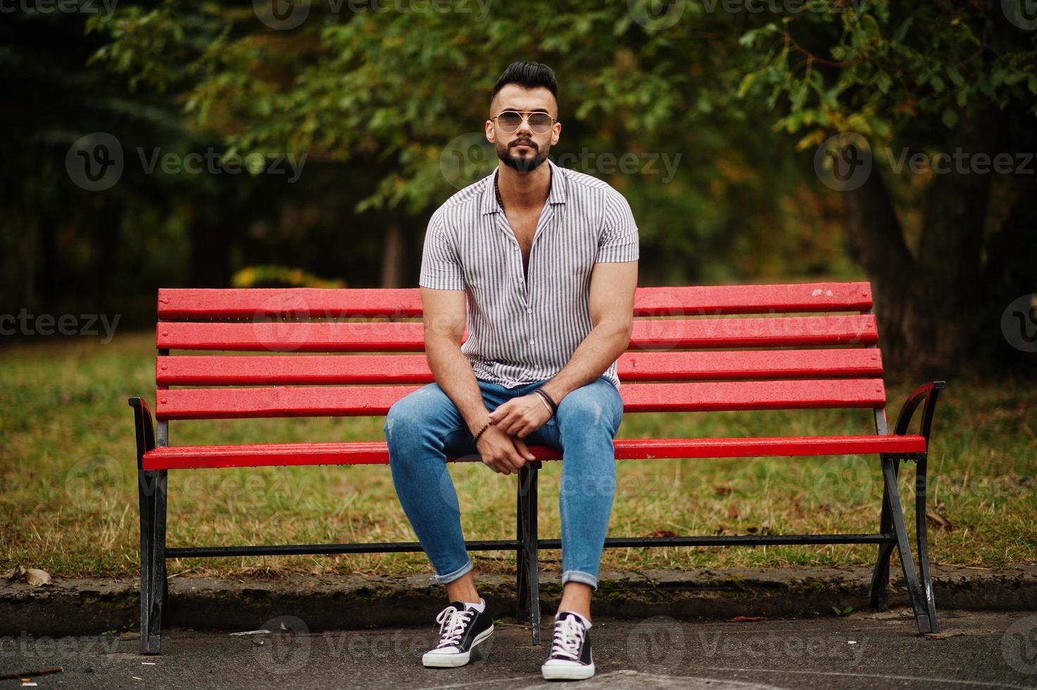 modieuze lange Arabische baard man slijtage op shirt, jeans en zonnebril zittend op een rode bank in het park. foto