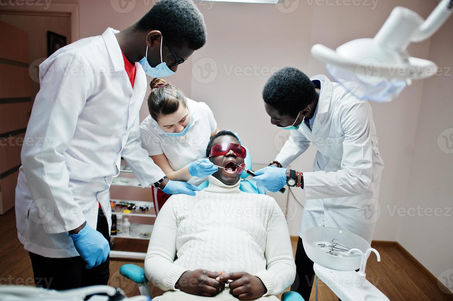 multiraciale tandarts artsen team. Afro-Amerikaanse man patiënt bij uv-beschermende bril. zijn tanden behandeld met behulp van een tandheelkundige uv-uithardingslamp en een tandheelkundige spiegel. foto