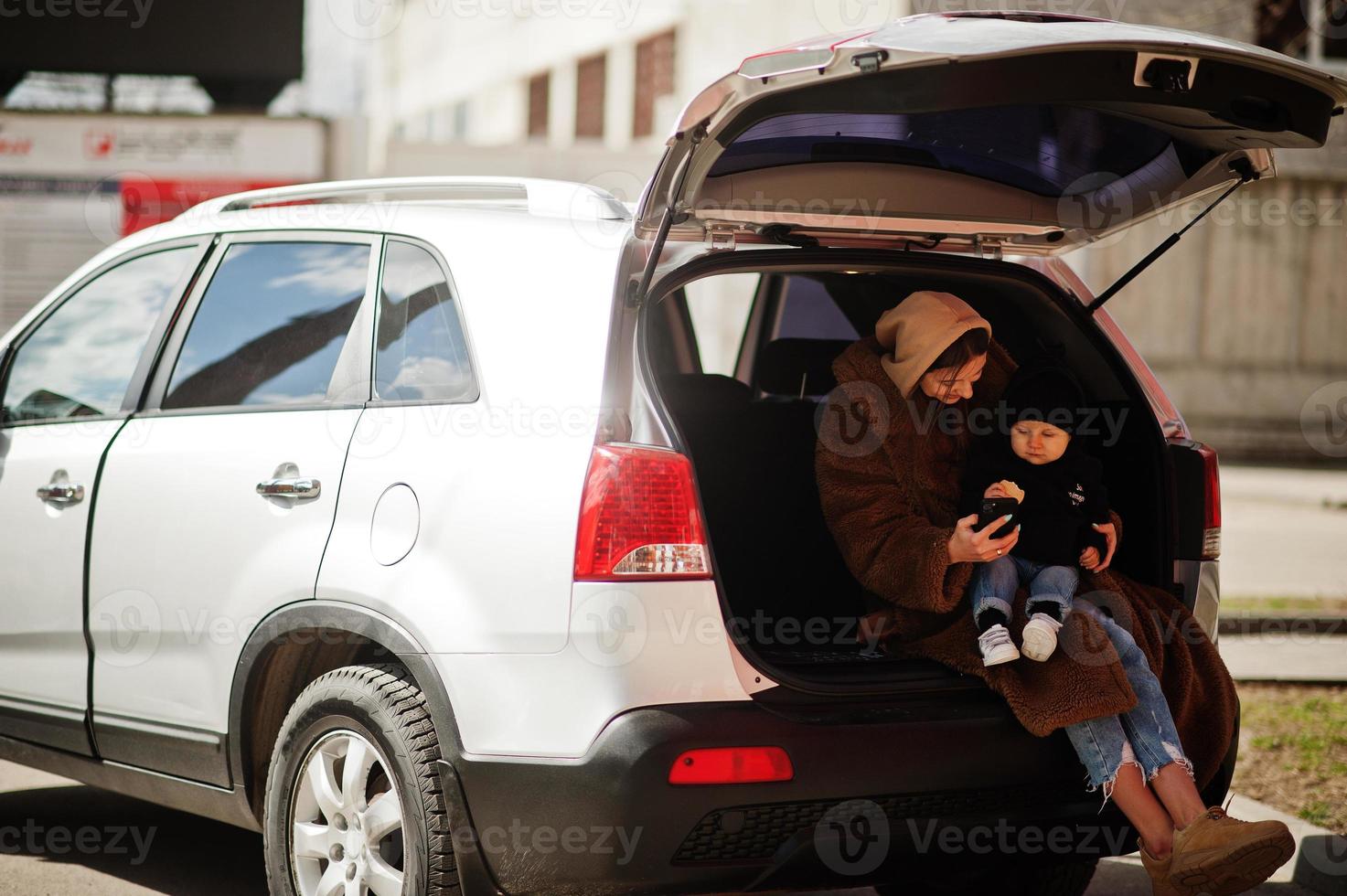 jonge moeder en kind zitten in de kofferbak van een auto en kijken naar mobiele telefoon. veiligheid rijden concept. foto