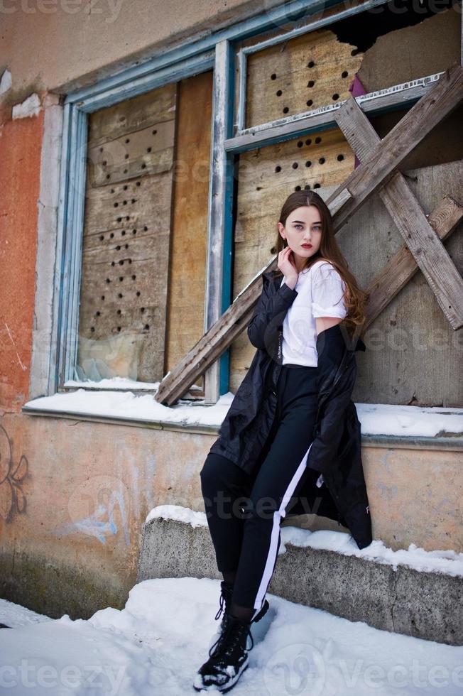 modieuze lange benen brunette model in lange zwarte mantel poseerde buiten op winterdag tegen oude grunge muur met gebroken ramen. foto