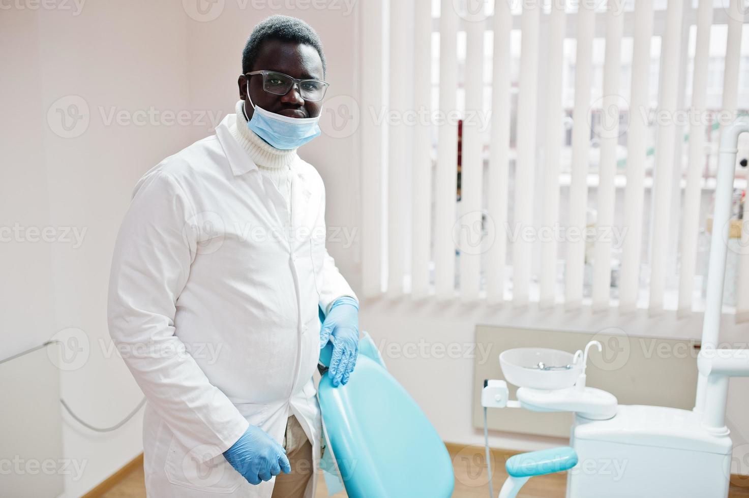 Afro-Amerikaanse mannelijke arts in glazen en masker staande in de buurt van tandartsstoel in tandheelkundige kliniek. foto