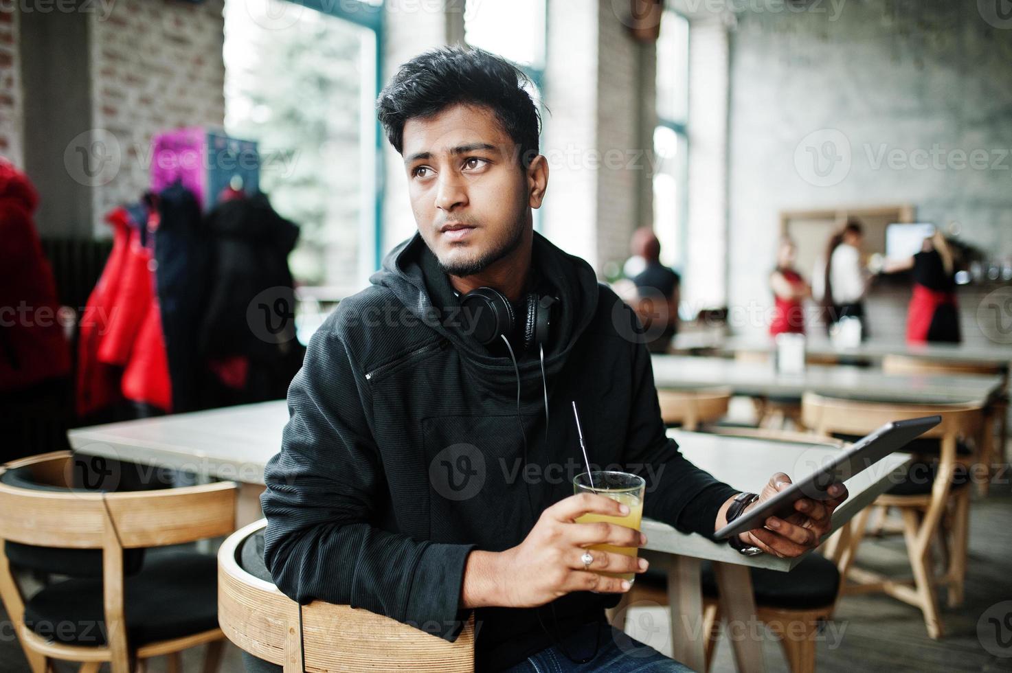 casual en stijlvolle jonge aziatische man met koptelefoon in café sap drinken uit glas en tablet vasthouden. foto