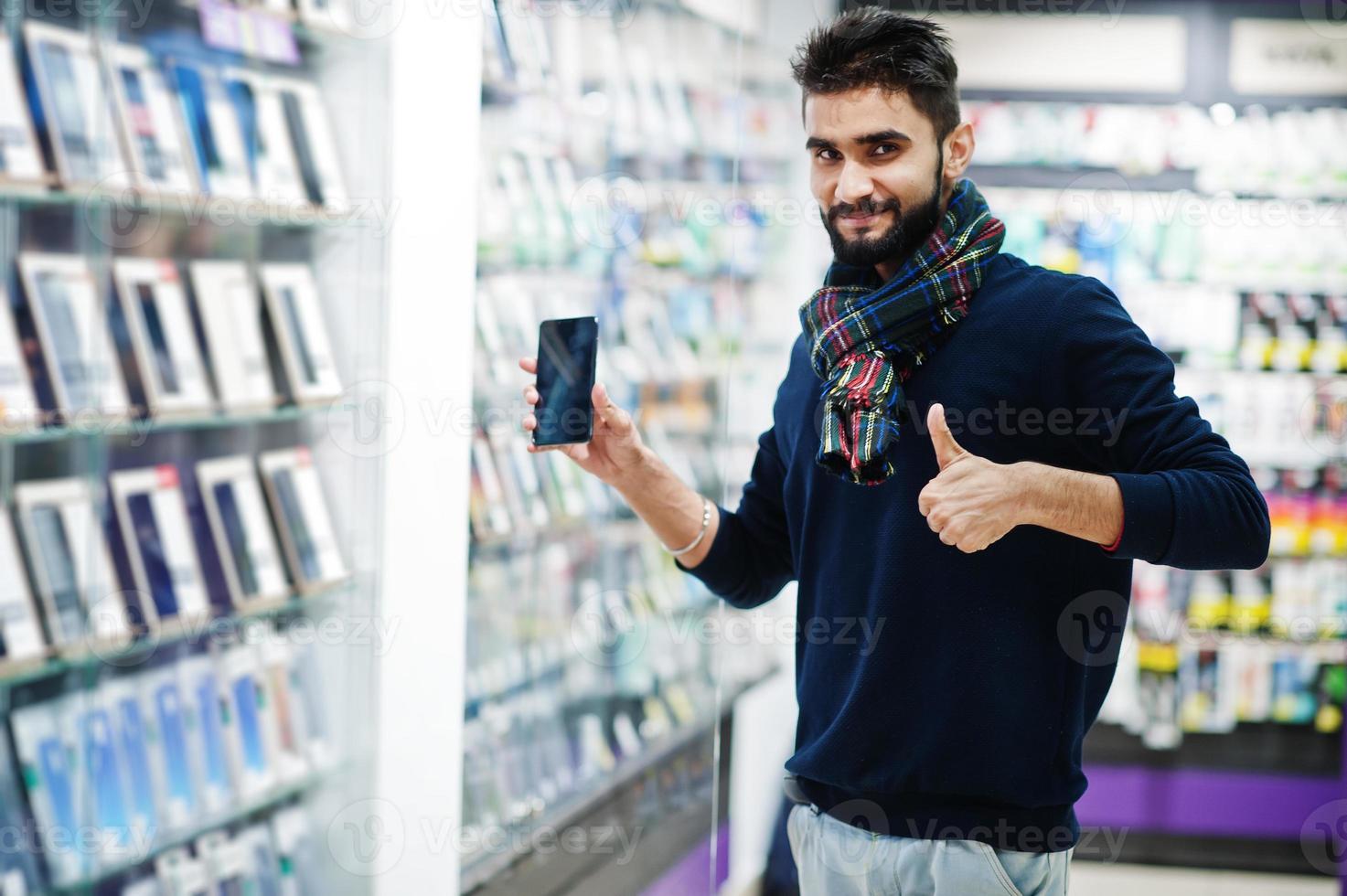 indiase baard man klant koper bij mobiele telefoon winkel kiest zijn nieuwe smartphone en toont duim. Zuid-Aziatische volkeren en technologieën concept. gsm winkel. foto