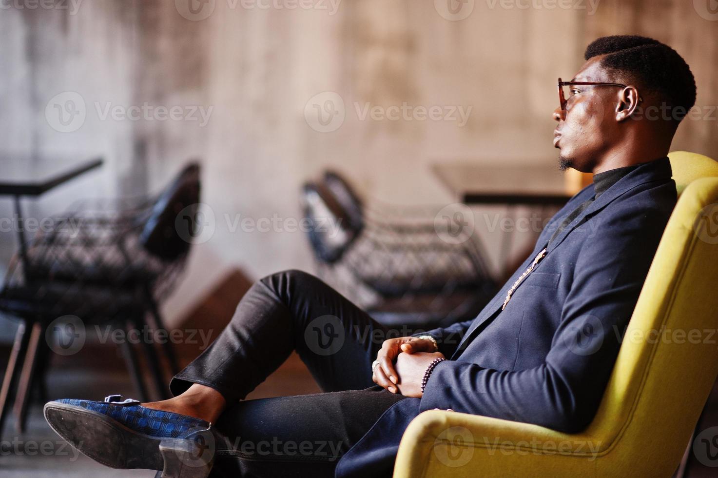 modieuze Afro-Amerikaanse man in pak en bril zittend op een stoel in café. foto