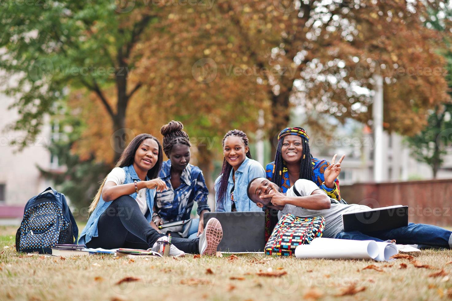 groep van vijf afrikaanse studenten die samen tijd doorbrengen op de campus op de universiteitswerf. zwarte afrovrienden die op gras zitten en met laptops studeren. foto