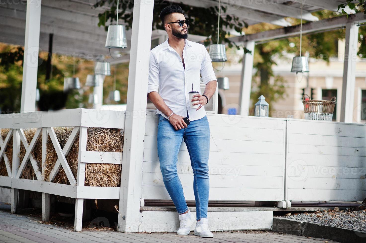stijlvolle lange Arabische man model in wit overhemd, jeans en zonnebril gesteld op straat van de stad. baard aantrekkelijke Arabische man met kopje koffie tegen terras. foto