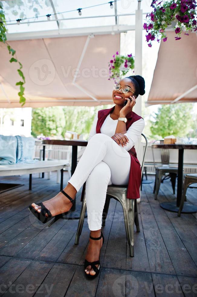 Afrikaans Amerikaans meisje draagt een bril met mobiele telefoon die aan de buitencaffe zit. foto