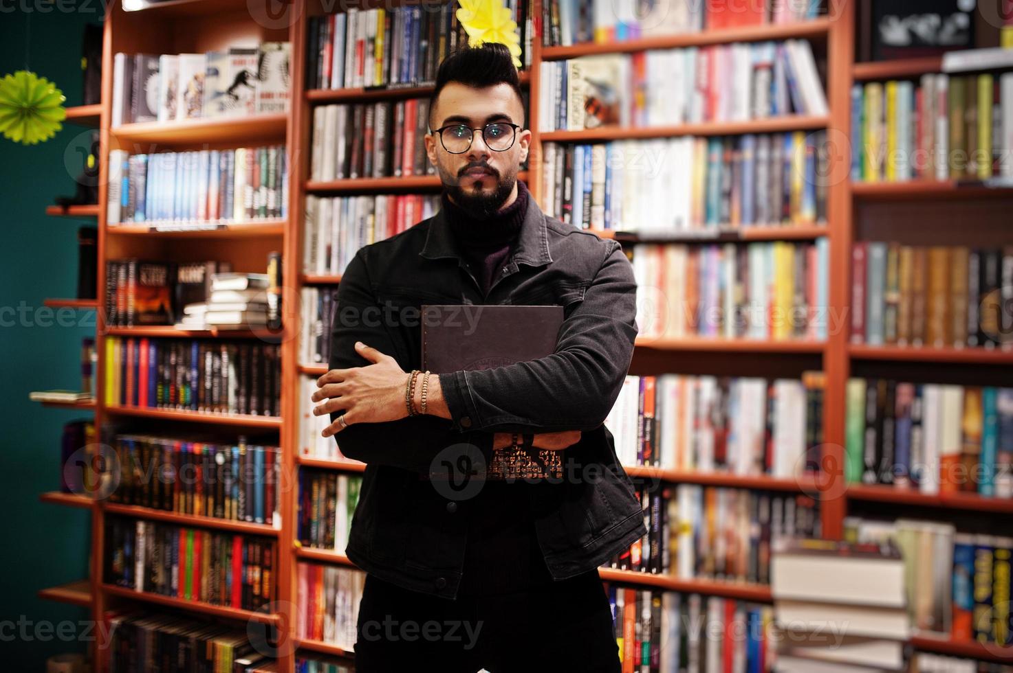 lange slimme arabische studentenman, draag een zwart spijkerjack en een bril, in de bibliotheek met een boek bij de hand. foto