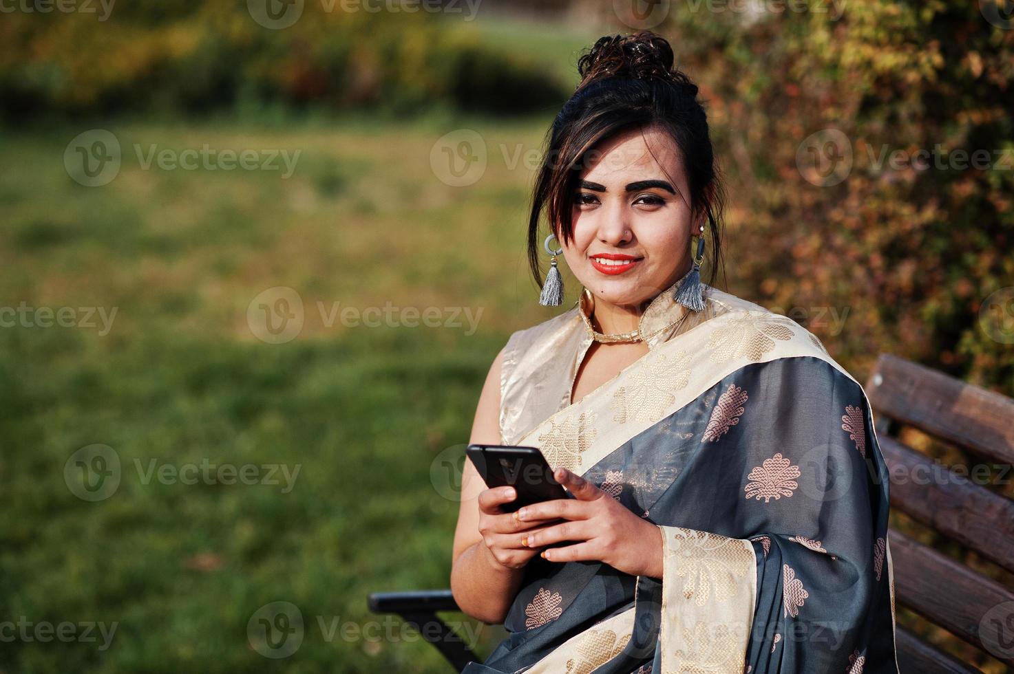 elegante brunette Zuid-Aziatische Indiase meisje in saree zittend op de bank met mobiele telefoon bij de hand. foto