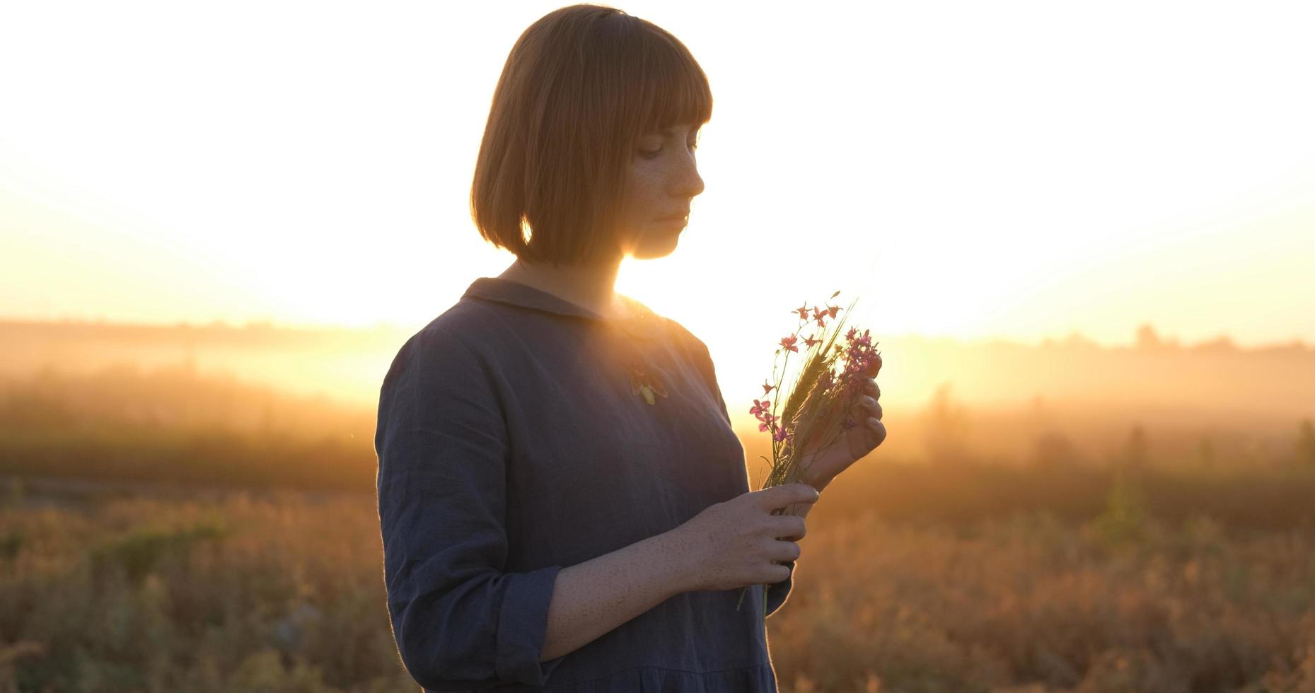 jong roodharige vrouw in mooi boho jurk ontspannende in de veld- gedurende mistig zonsondergang, vrouw buitenshuis met boeket in handen foto