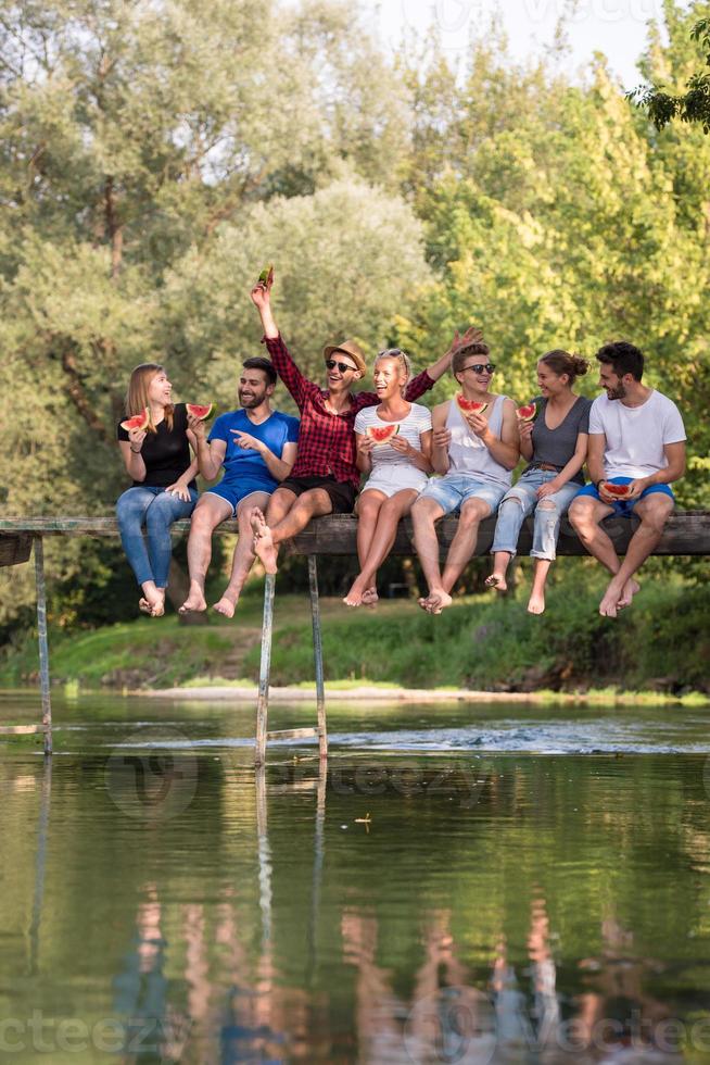 vrienden genieten van watermeloen terwijl zittend Aan de houten brug foto