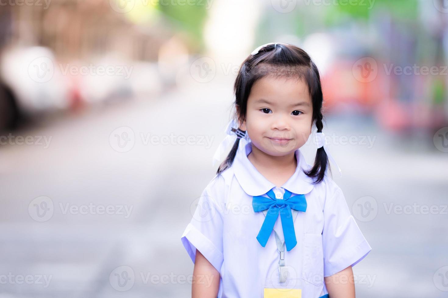 leerling stond glimlachen zoet op zoek Bij de camera. kind staan en wacht voor moeder voordat gaan naar school. schattig Aziatisch kind meisje vervelend school- uniform, blauw en wit, 3 jaren oud. gevlochten vlecht. foto