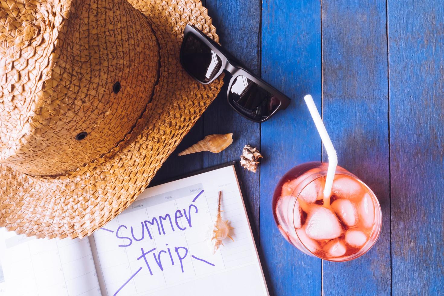 hoed met zonnebril, rood cocktail, kalender en schelpen Aan blauw geschilderd hout plank achtergrond. zomer vakantie concept foto