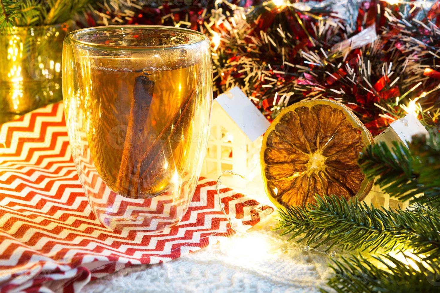 transparant dubbelwandig glas tuimelaar met heet thee en kaneel stokjes Aan de tafel met Kerstmis decor en klein huis. nieuw jaar atmosfeer, plak van droog oranje, guirlande, net tak, knus foto