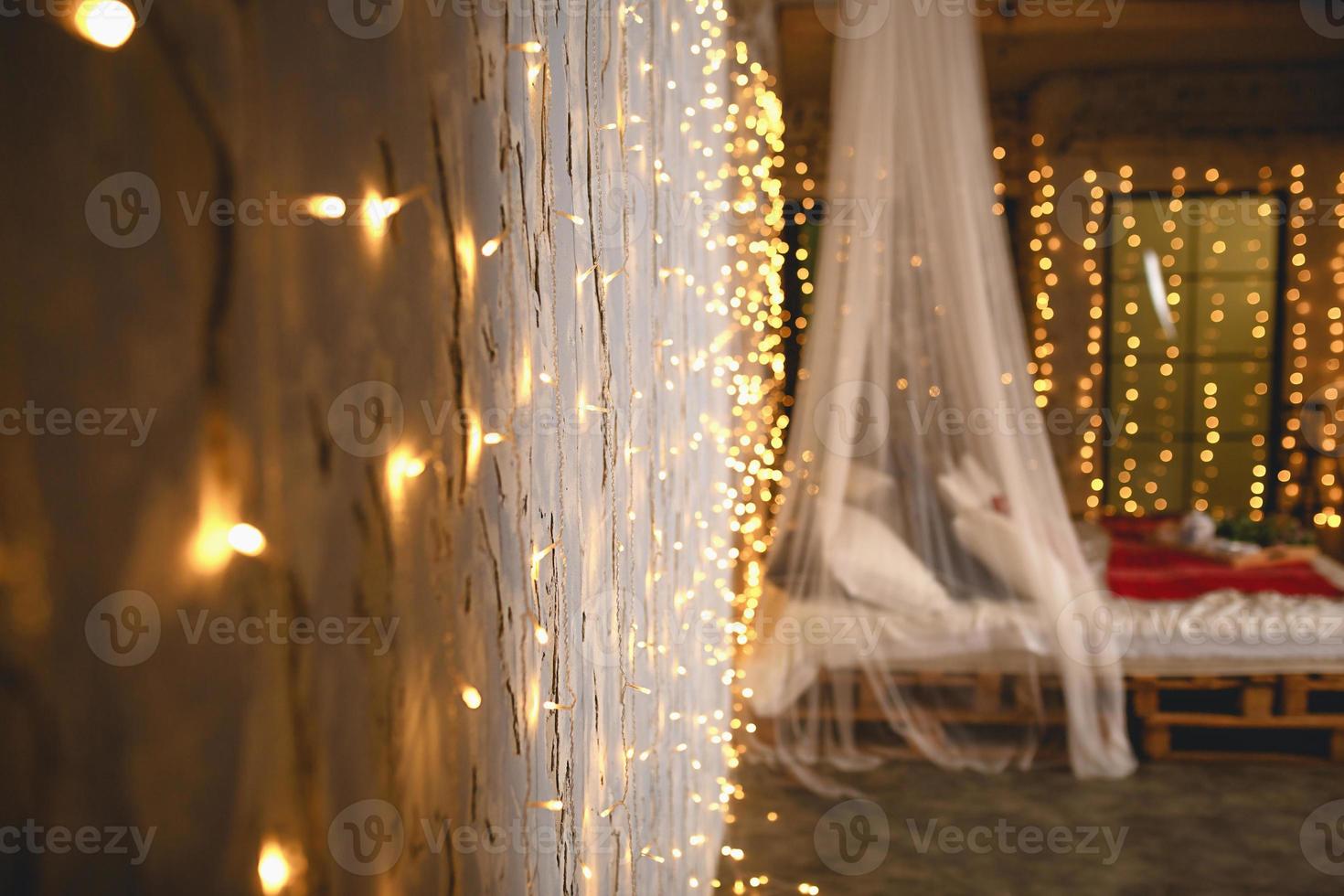 Kerstmis decor in de slaapkamer in de zolder stijl. een hemelbed bed gemaakt van houten pallets met wit linnen en rood dekens en kussens. net en goud slingers, verlichte venster. nieuw jaar. foto