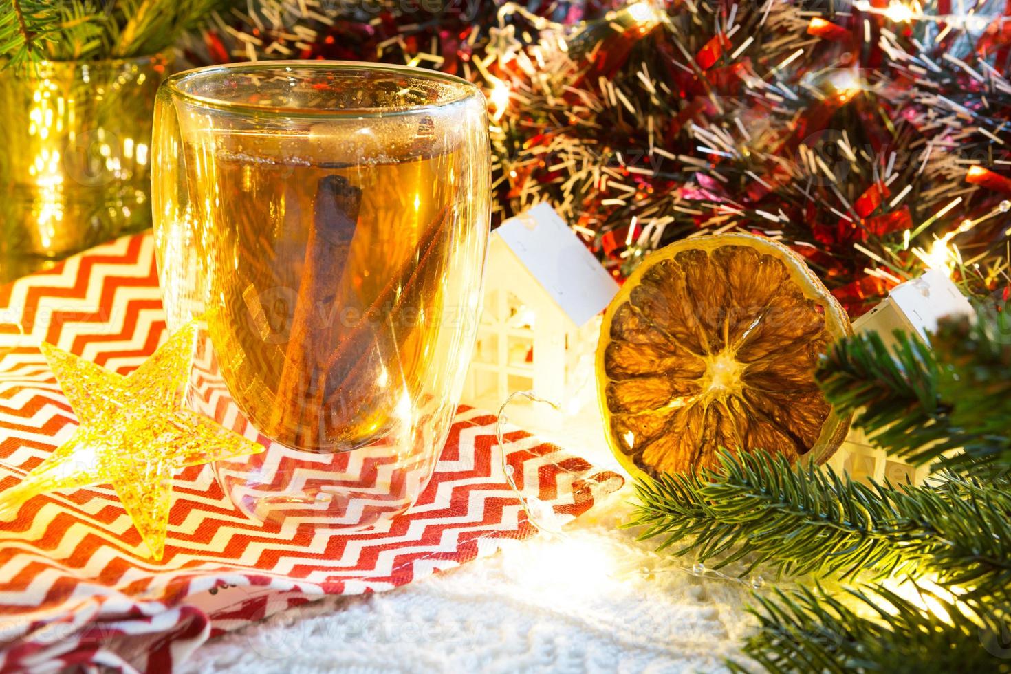 transparant dubbelwandig glas tuimelaar met heet thee en kaneel stokjes Aan de tafel met Kerstmis decor en klein huis. nieuw jaar atmosfeer, plak van droog oranje, guirlande, net tak, knus foto