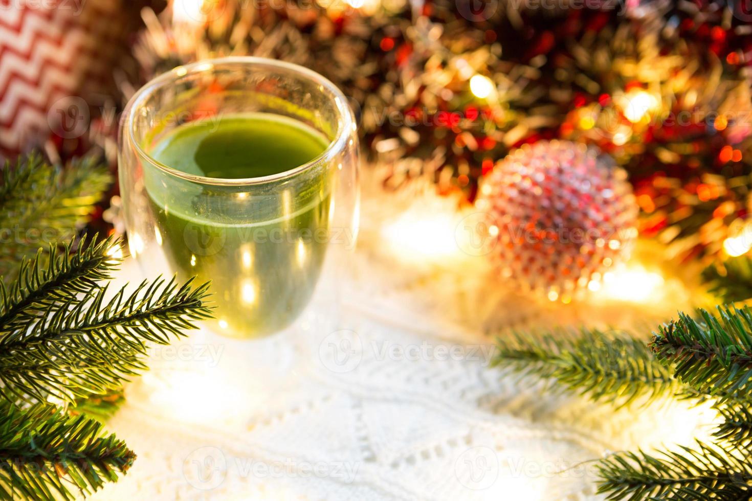 transparant dubbelwandig glas tuimelaar met Japans matcha thee Aan tafel met Kerstmis decor. nieuw jaar atmosfeer, slinger en klatergoud, net tak, knus, gebreid deken, bal, gestreept servet foto