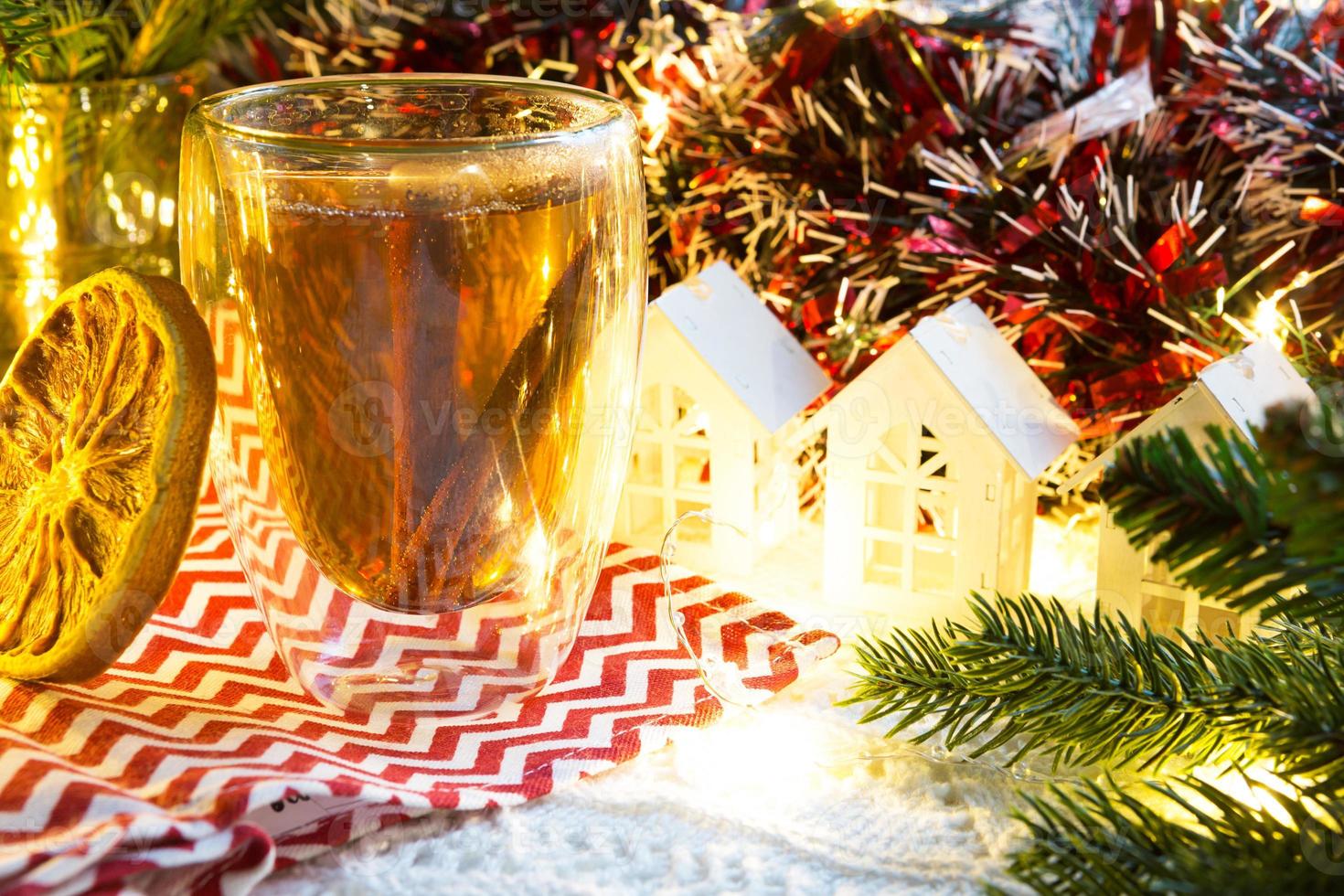 transparant dubbelwandig glas tuimelaar met heet thee en kaneel stokjes Aan de tafel met Kerstmis decor en klein huis. nieuw jaar atmosfeer, plak van droog oranje, guirlande, net tak, knus foto