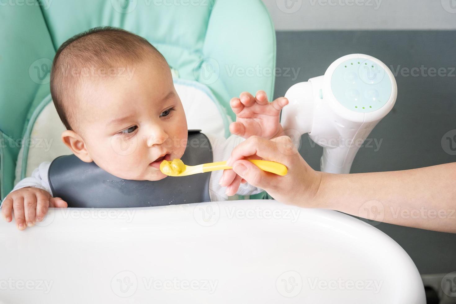 mam feeds de baby met een lepel van groente puree Bij de kinderen voeden tafel. baby's trek, gezond voeding, invoering van complementair voedingsmiddelen. kopieerruimte, bespotten omhoog foto