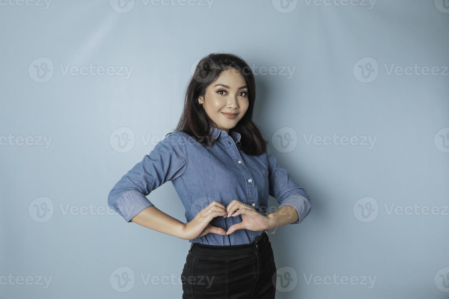 een aantrekkelijk jong Aziatisch vrouw vervelend een blauw overhemd voelt gelukkig en een romantisch vormen hart gebaar drukt uit inschrijving gevoelens foto