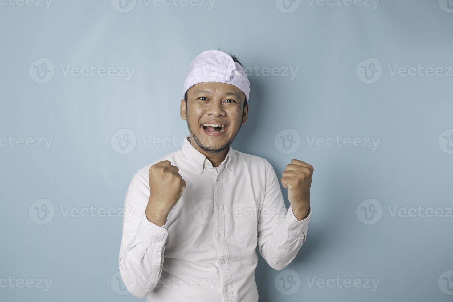 een jong balinees Mens met een gelukkig geslaagd uitdrukking vervelend udeng of traditioneel hoofdband en wit overhemd geïsoleerd door blauw achtergrond foto