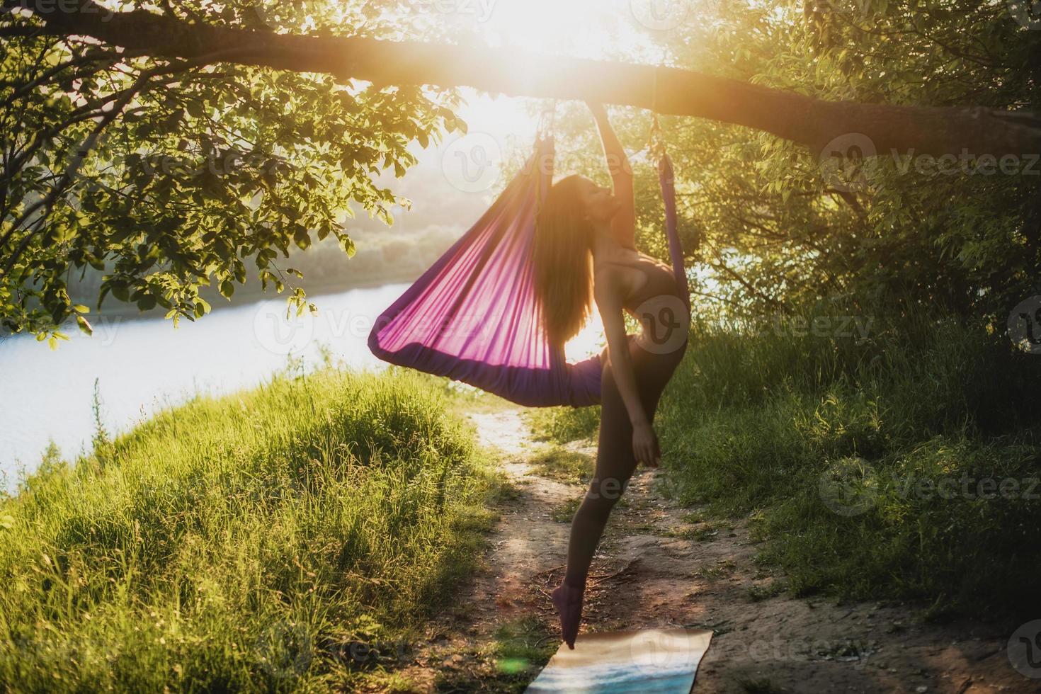 een jong vrouw gymnast is verloofd in antenne yoga, gebruik makend van een combinatie van traditioneel yoga poseert, pilates en dans gebruik makend van een hangmat Bij zonsondergang in natuur. gezond levensstijl. foto