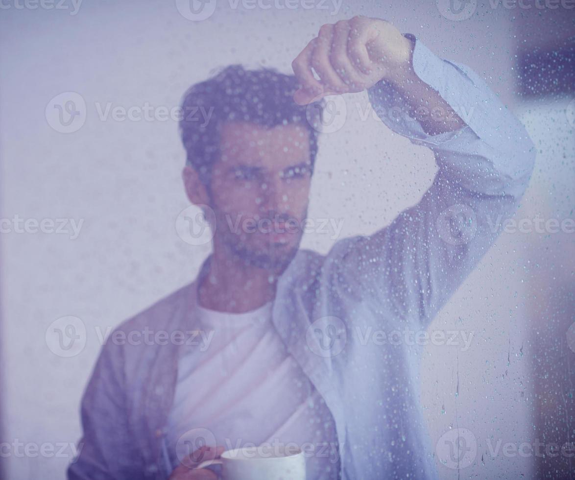 ontspannen jong Mens drinken eerste ochtend- koffie meth regen druppels Aan venster foto