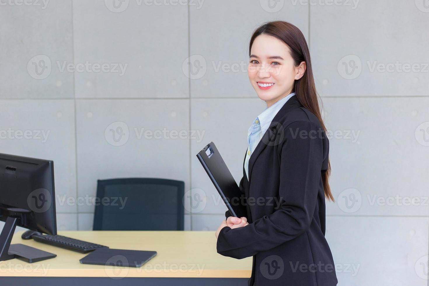 jonge Aziatische professionele werkende vrouw in een zwart pak houdt klembord in haar handen en zelfverzekerde glimlach in kantoorruimte. foto