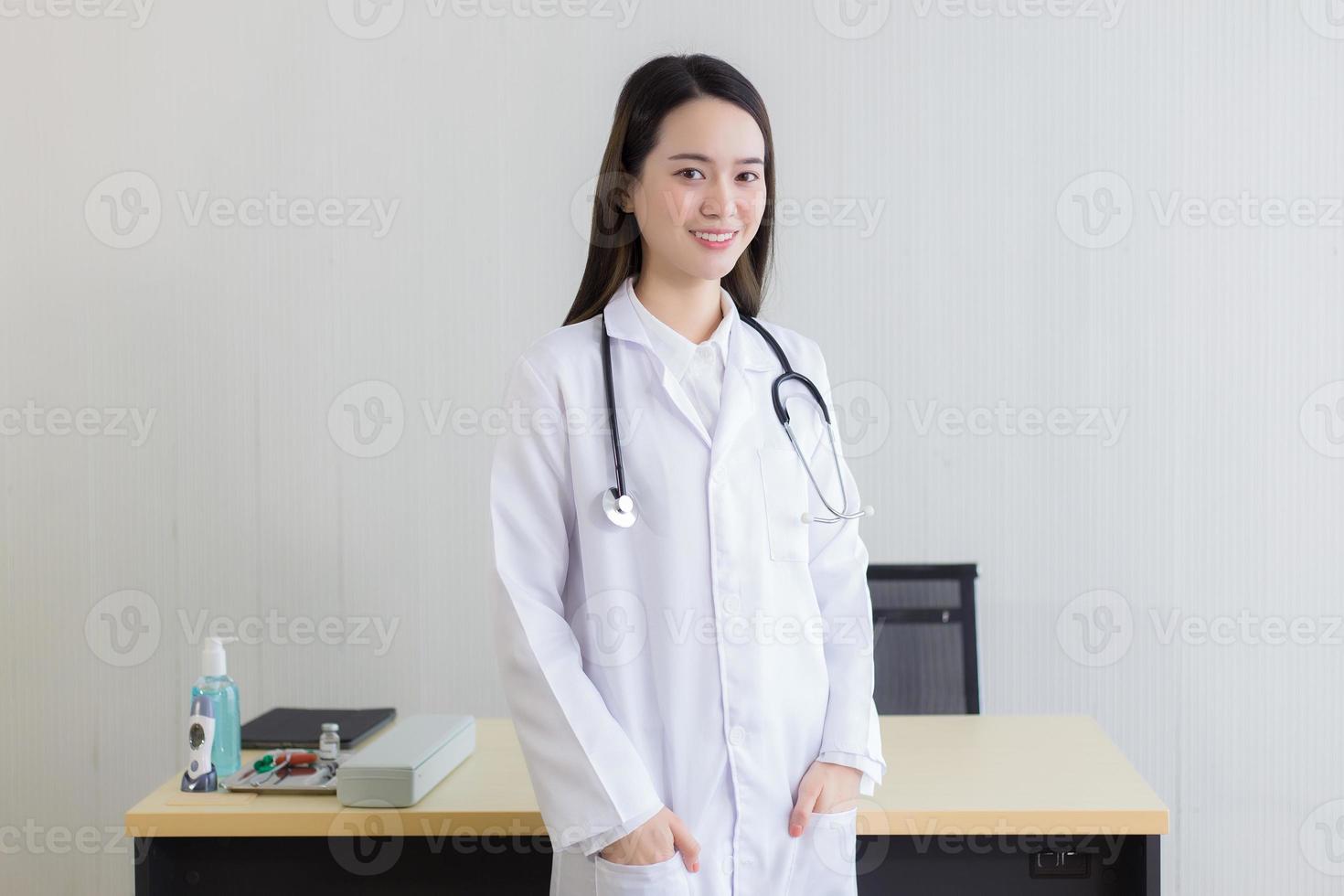 een aziatische vrouwelijke arts met een witte laboratoriumjas die staat en een wit gewaad en een stethoscoop draagt. foto