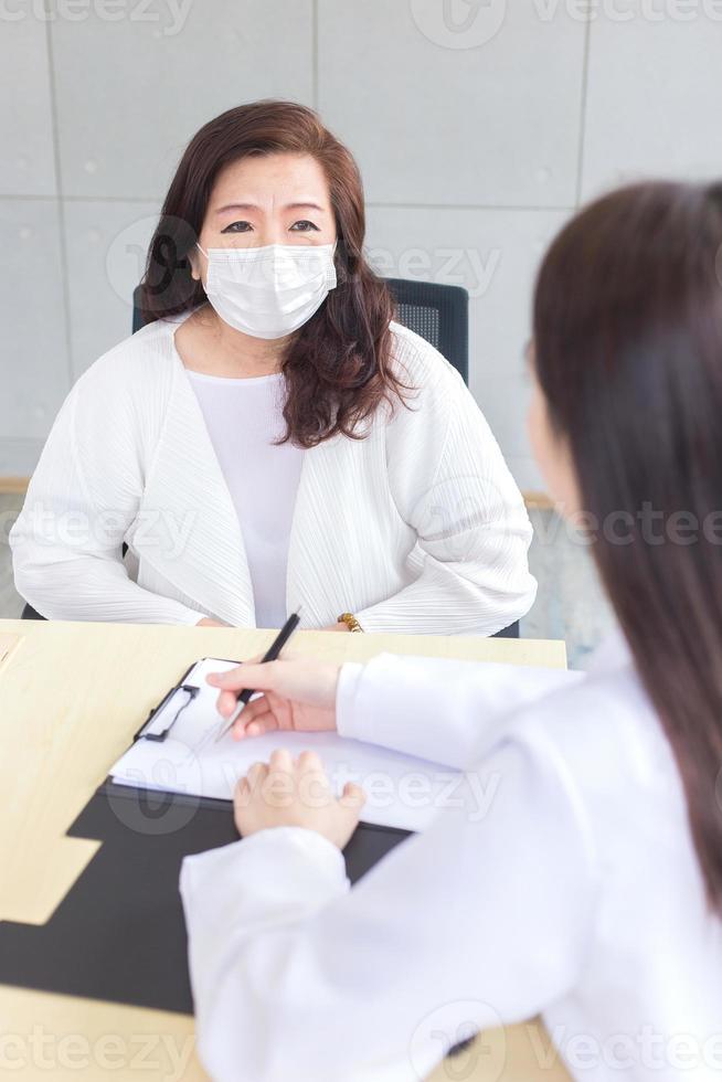 Aziatisch vrouw dokter records symptoom van geduldig Aan de document terwijl diagnose, beide slijtage gezicht masker in ziekenhuis. foto