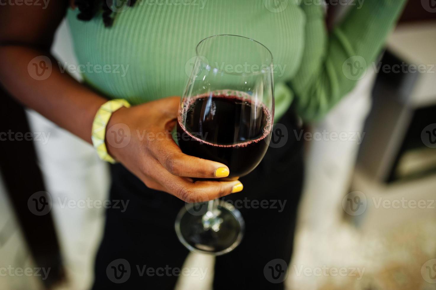 dichtbij omhoog handen van aantrekkelijk jong Afrikaanse Amerikaans vrouw vervelend in groen trui en zwart jeans houding Bij restaurant met glas van rood wijn. foto