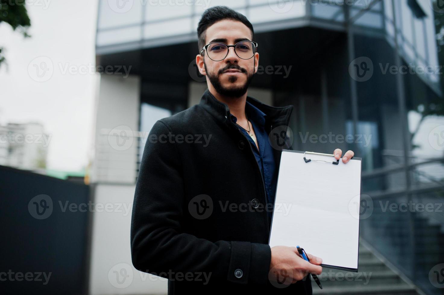 midden- oostelijk Mens slijtage zwart jas, bril, houden wit leeg klembord. foto