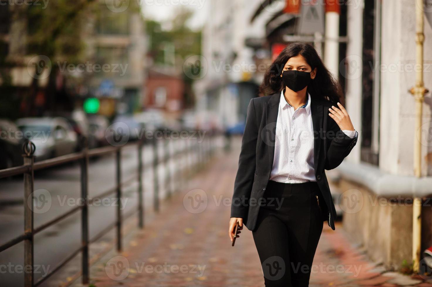 prachtig Indisch vrouw slijtage formeel en zwart gezicht masker, poseren Bij straat gedurende covid pandemie, met mobiele telefoon Bij hand. foto