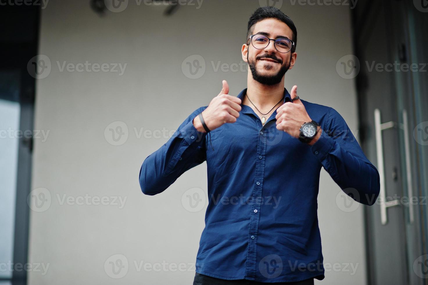 midden- oostelijk ondernemer slijtage blauw shirt, bril tegen kantoor gebouw en tonen duimen omhoog. foto