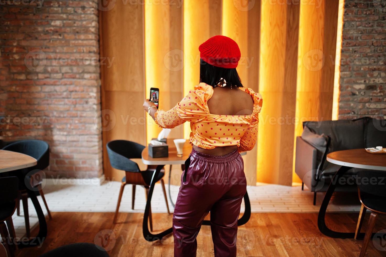 elegant afro Amerikaans vrouw in rood Frans baret, groot goud nek keten polka punt blouse en leer broek voert een leven uitzending door mobiel telefoon voor sociaal netwerk. foto