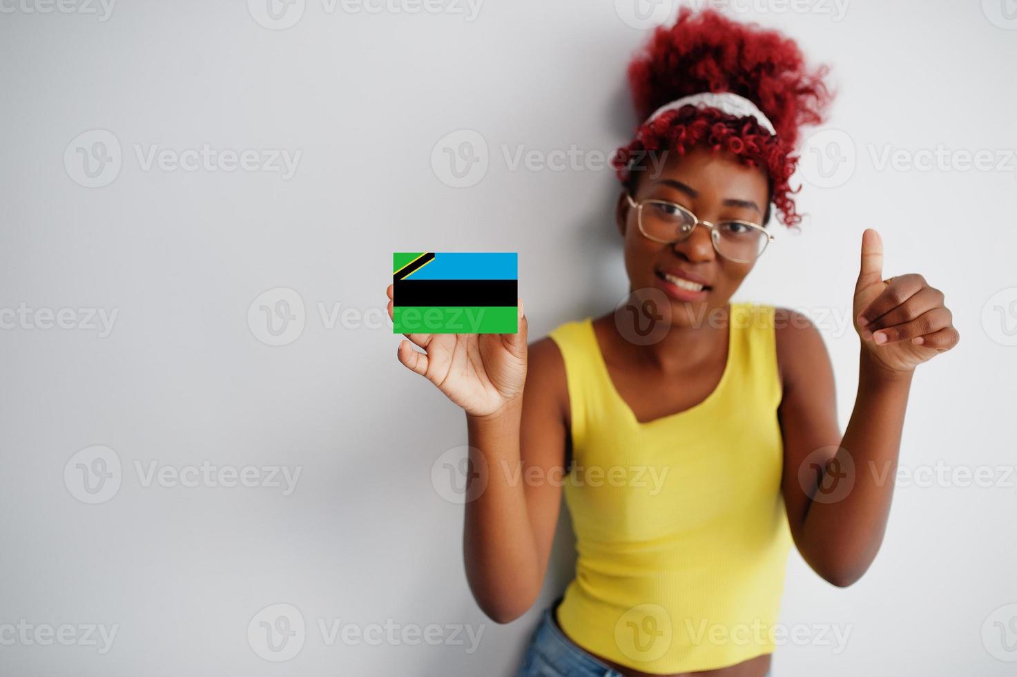 Afrikaanse vrouw met afro haar, slijtage geel singlet en bril, houden zanzibar vlag geïsoleerd Aan wit achtergrond, tonen duim omhoog. foto