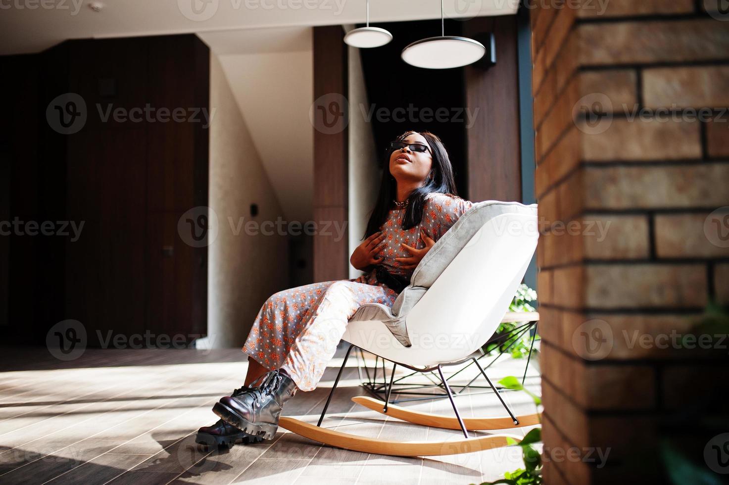 mode portret van Afrikaanse Amerikaans vrouw in lang jurk, laarzen en zonnebril binnen. foto