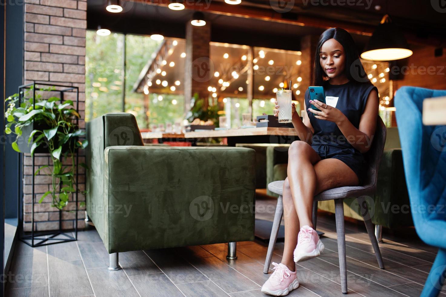 modieus feministisch Afrikaanse Amerikaans vrouw slijtage in zwart t-shirt en korte broek, gesteld Bij restaurant met limonade glas en mobiel telefoon. foto