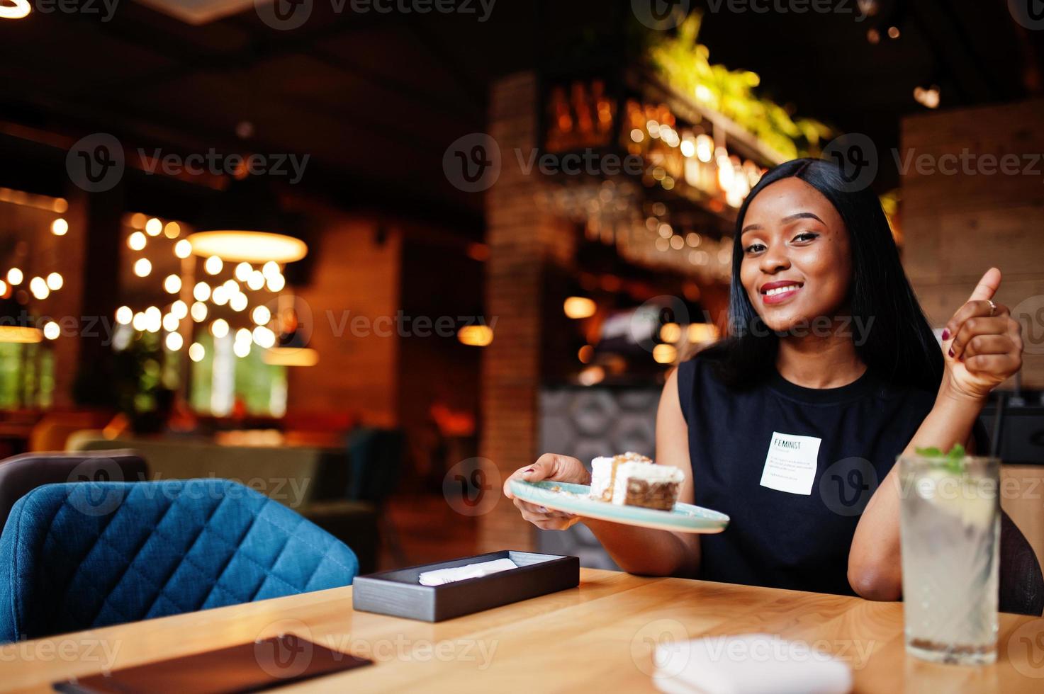 modieus feministisch Afrikaanse Amerikaans vrouw slijtage in zwart t-shirt en korte broek, gesteld Bij restaurant, eten kaas taart. foto