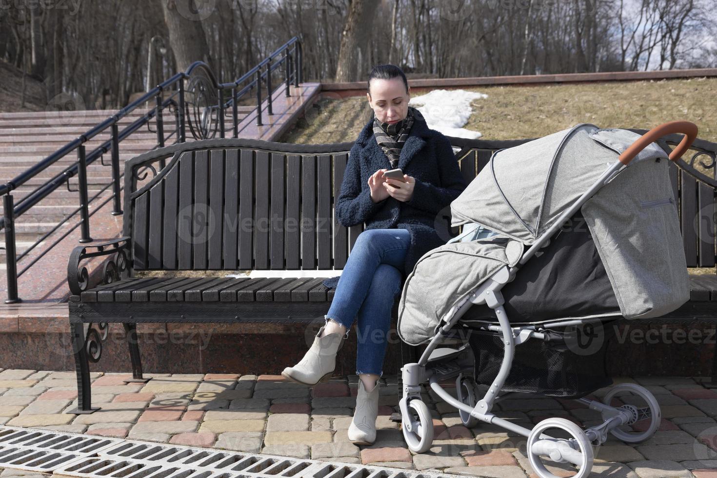 jong vrouw wandelingen in de park in de voorjaar met een baby koets. moeder looks in een mobiel telefoon terwijl de baby slaapt buitenshuis. foto