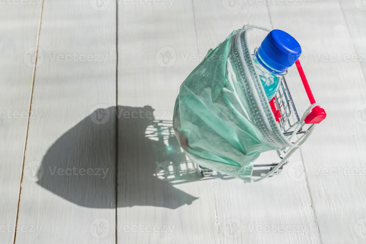 boodschappen doen kar in medisch masker met fles van ontsmettingsmiddel Aan wgite houten achtergrond. coronavirus het voorkomen concept. top visie. foto