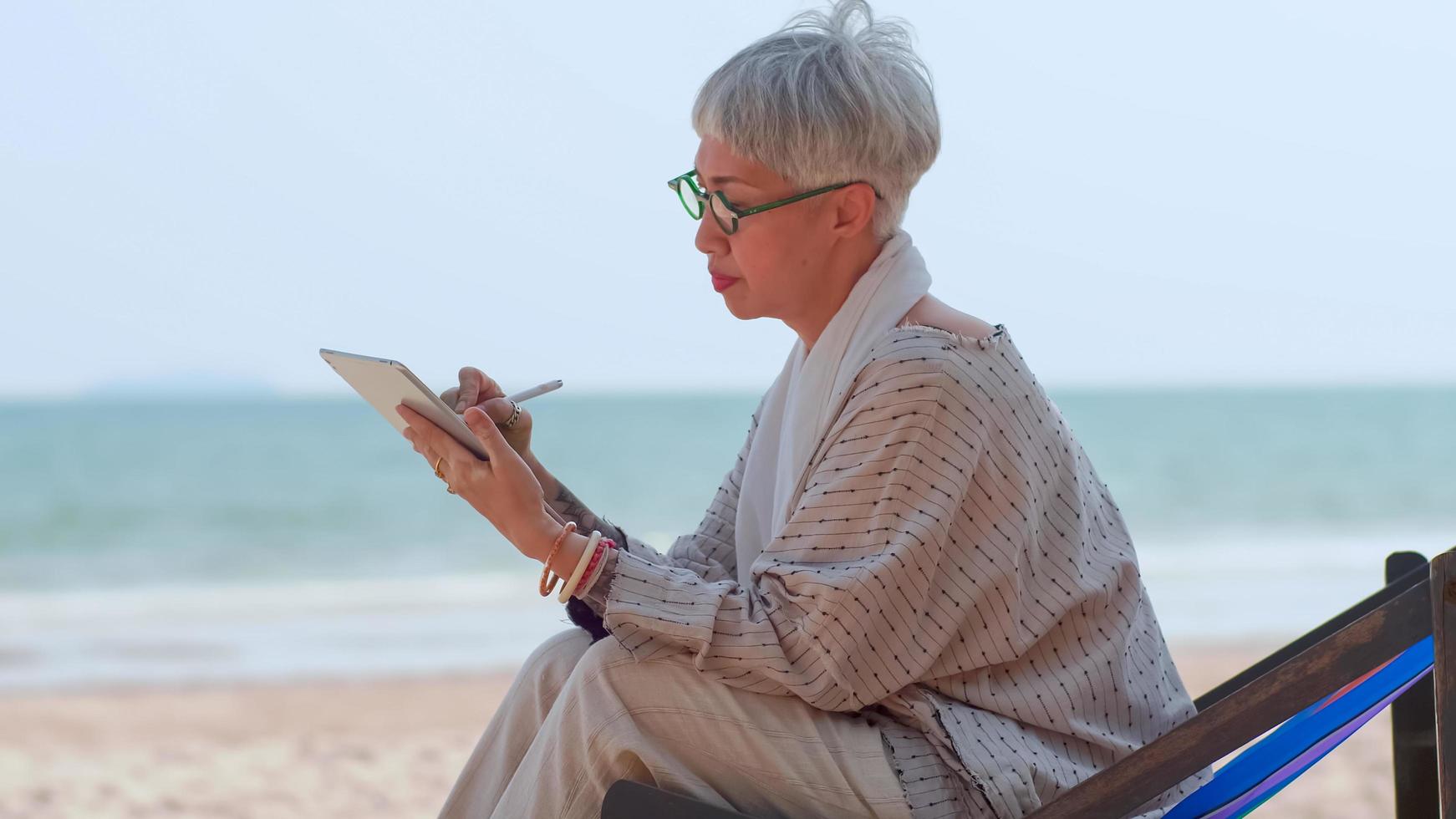 Aziatisch ouderen vrouw gebruik makend van een tablet computer terwijl zittend Aan de strand. foto