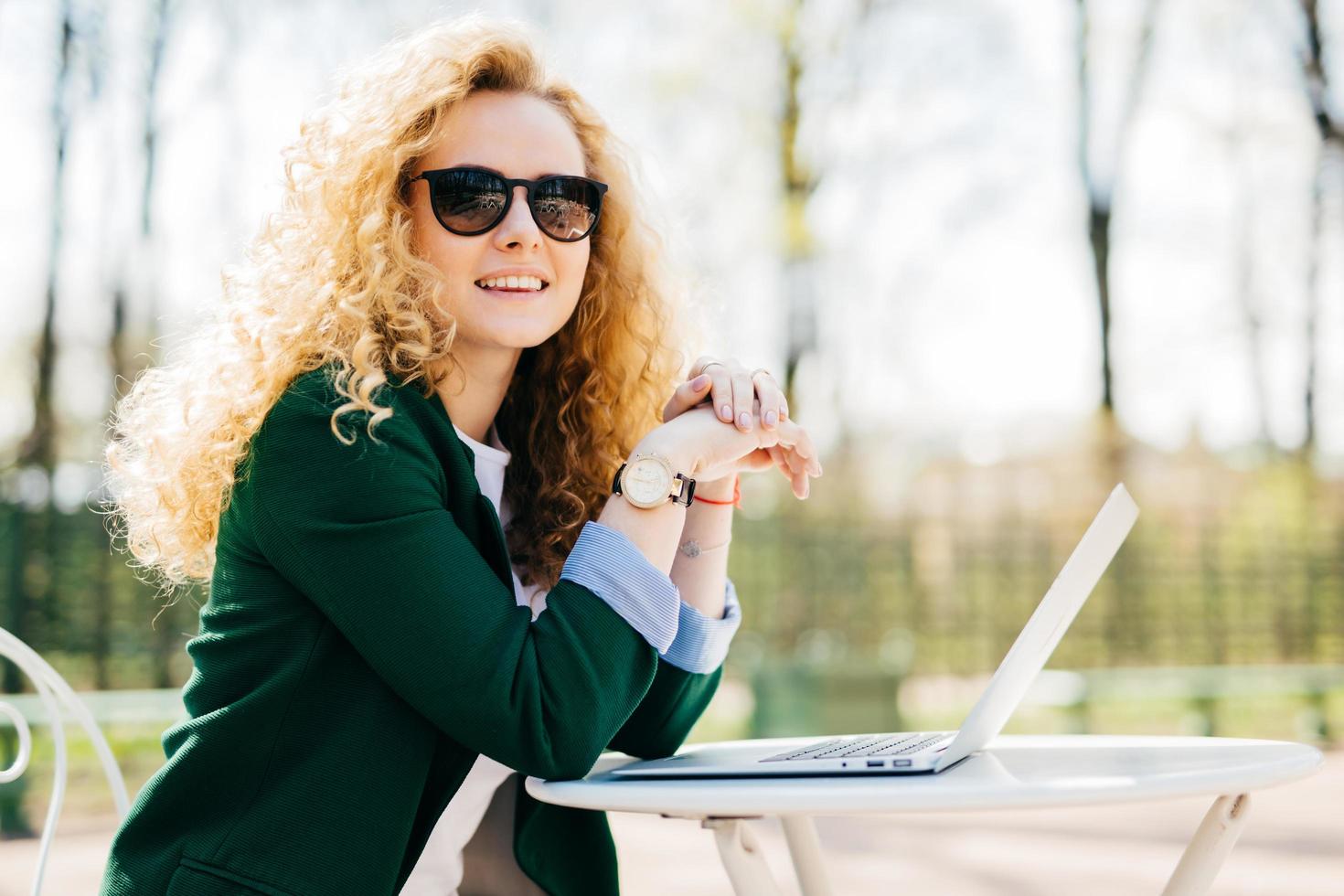 mooie jonge studente vrouw met mooi krullend blond haar elegant gekleed met behulp van generieke laptopcomputer voor het schrijven van essay met rust buitenshuis met charmante glimlach op zoek naar afstand foto