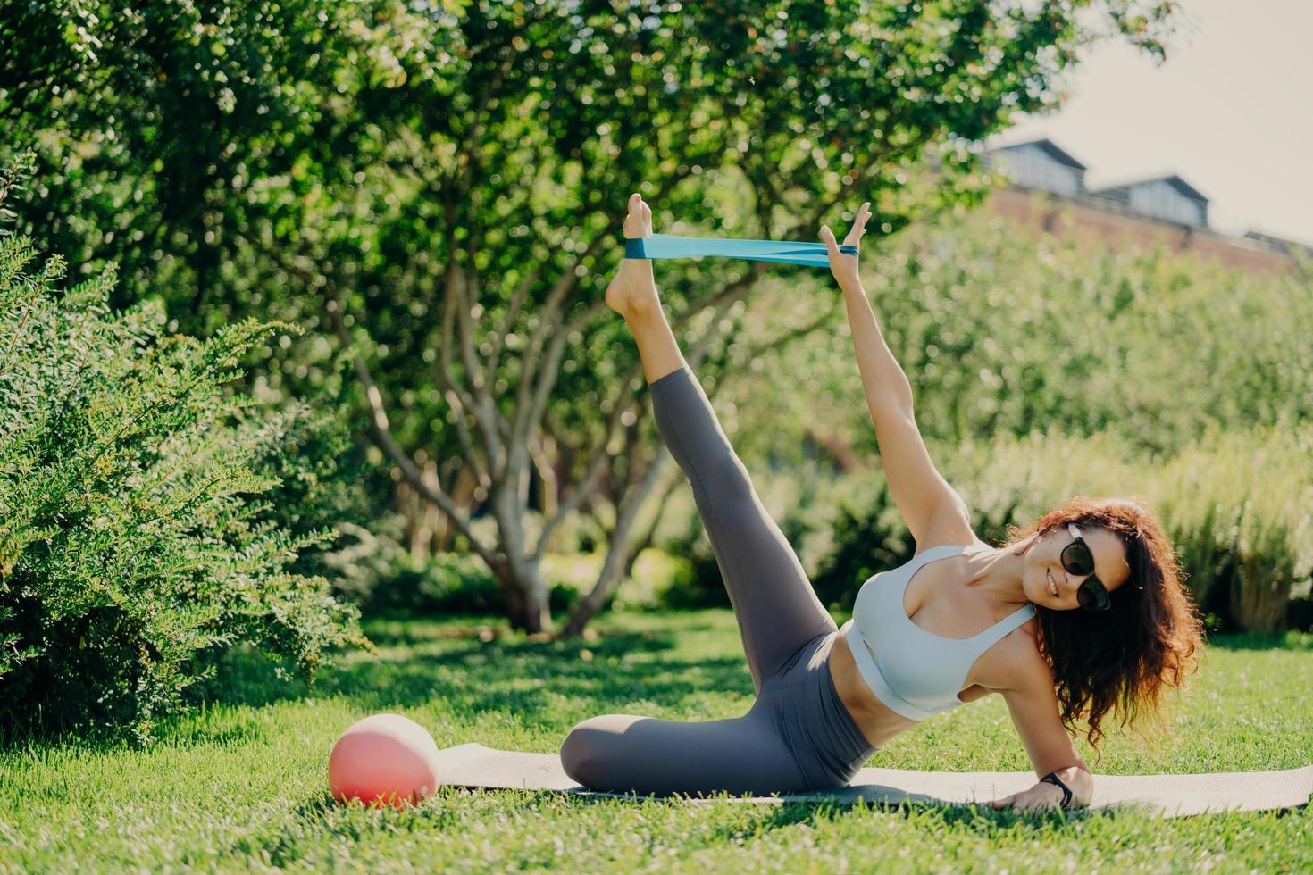 sterke fit vrouw gebruikt fitnessgom om benen en armen te strekken staat op zijplank bij karemat gekleed in sportkleding poses buiten tijdens zomerdag alles is groen. oefeningen op beenspieren foto