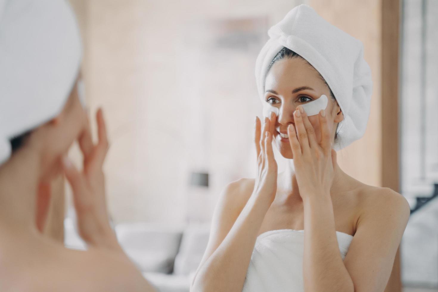 meisje past ooglapjes toe en kijkt in de spiegel. aantrekkelijke vrouw gewikkeld in een handdoek na het baden. foto