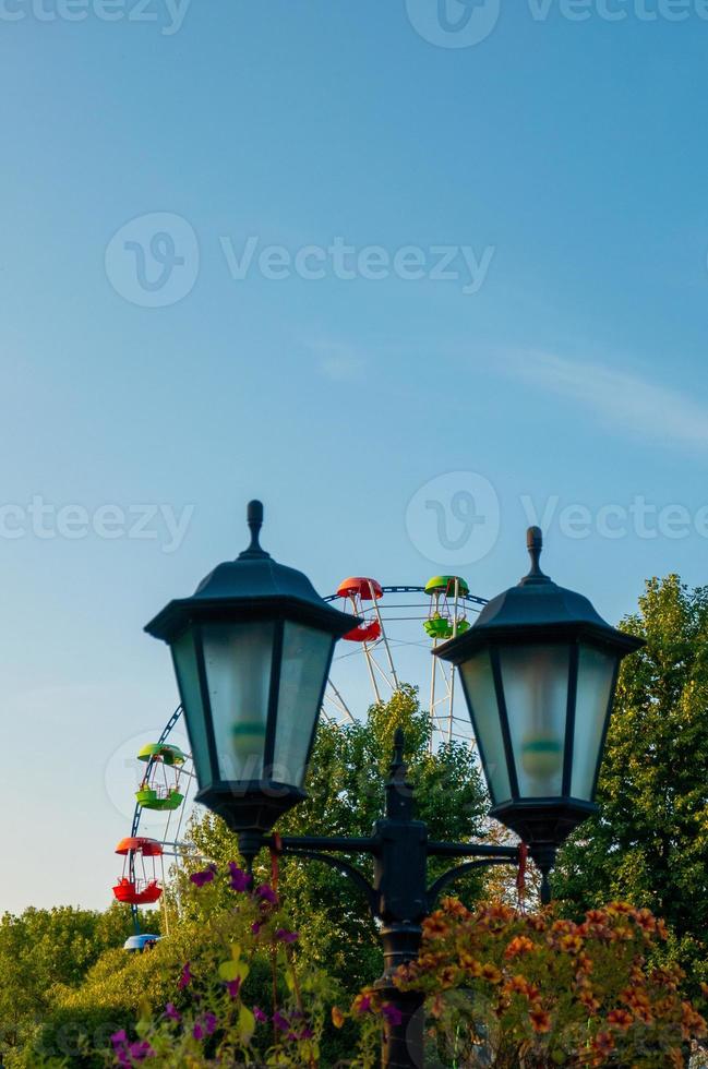 landschap van een pretpark met lantaarn versierd met bloemen achtergrond de top van een reuzenrad met boven de boomtoppen tegen een blauwe hemel. foto