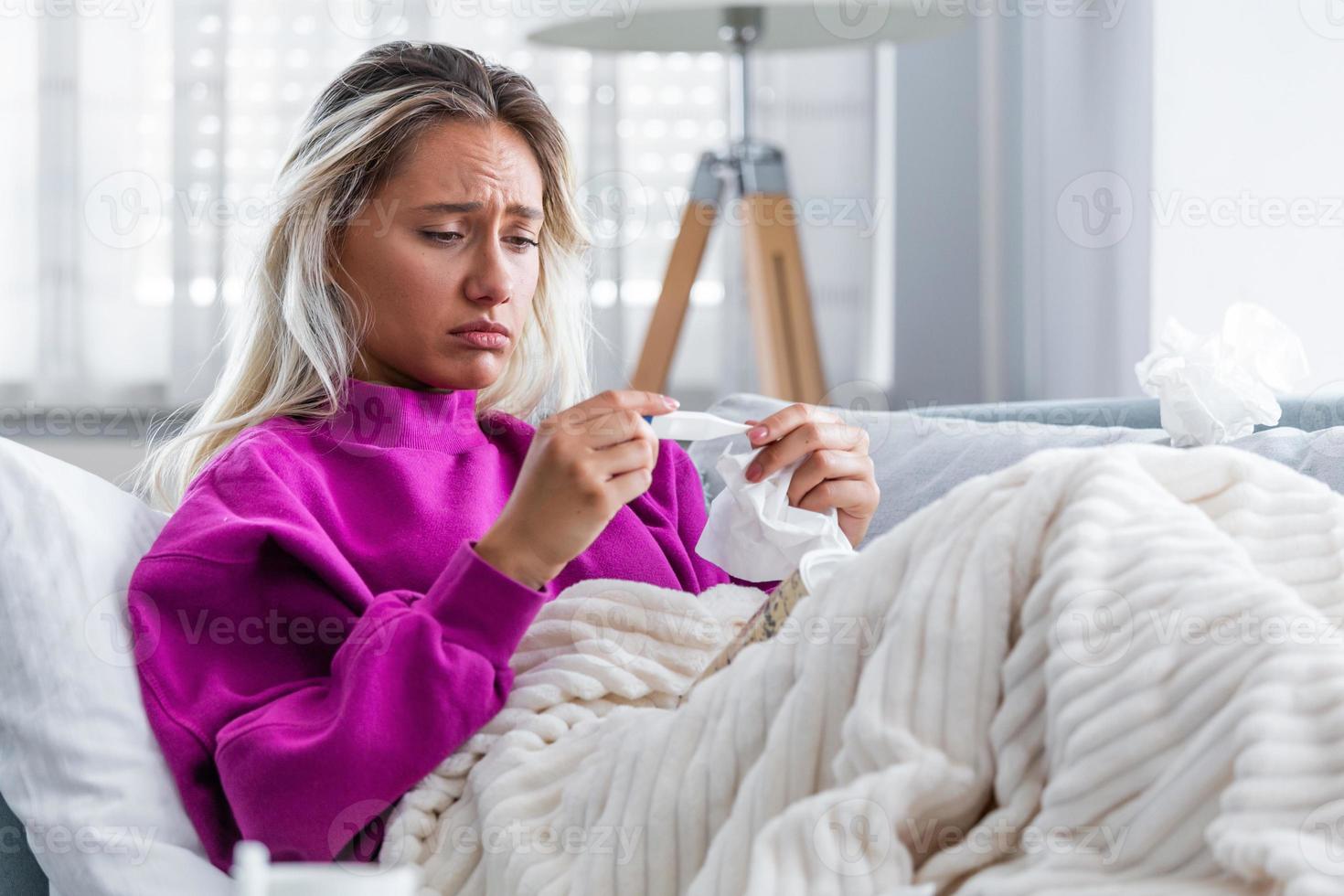 ziekte, seizoensgebonden virus probleem concept. vrouw wezen ziek hebben griep aan het liegen Aan sofa op zoek Bij temperatuur Aan thermometer. ziek vrouw aan het liegen in bed met hoog koorts. verkoudheid griep en migraine. foto